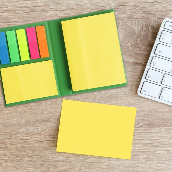A compact sticky notepad with yellow pages and colourful sticky flags, placed on a wooden surface beside a keyboard.