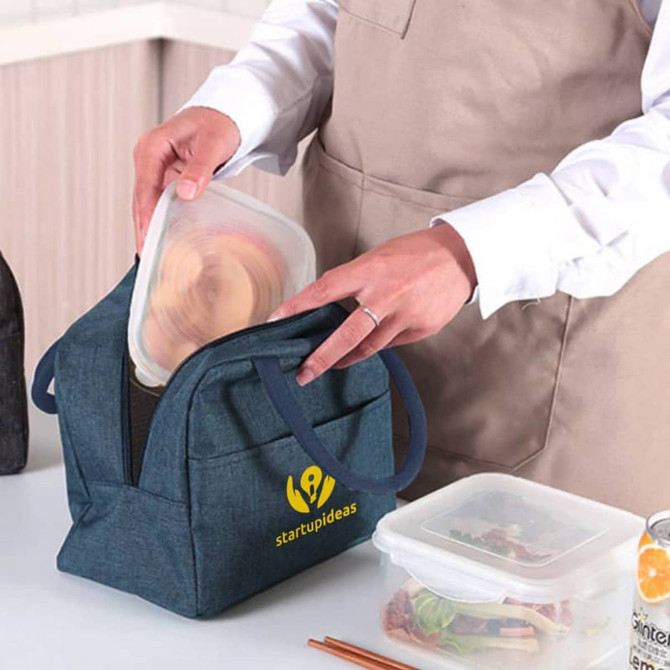 A denim blue cooler lunch bag with handles, featuring a logo, and a person placing a container inside.