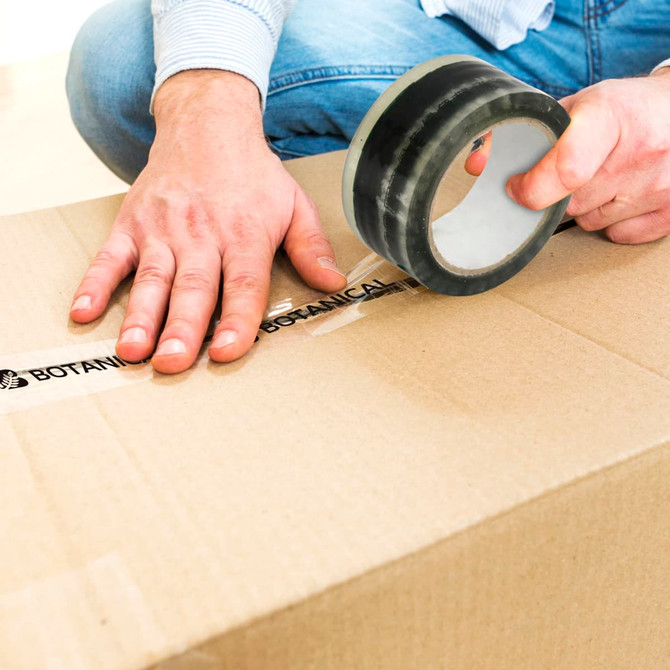 A person sealing a brown cardboard box with black BOPP packing tape, while wearing blue jeans and a light-coloured shirt.
