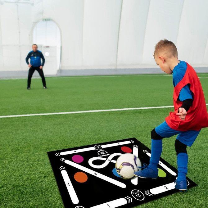 A black football training mat with white lines and colourful circles, placed on green turf, with a player and coach visible.