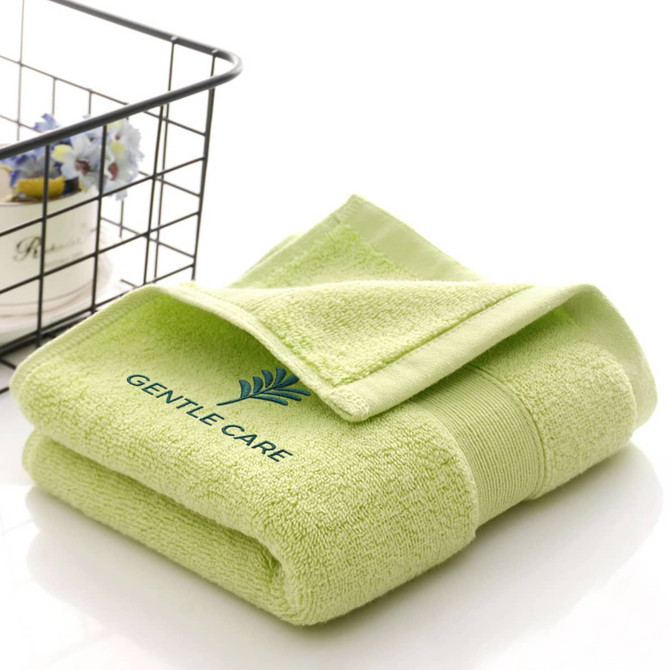 A folded green embroidered cotton hand towel displayed next to a wire basket. The towel features a logo.