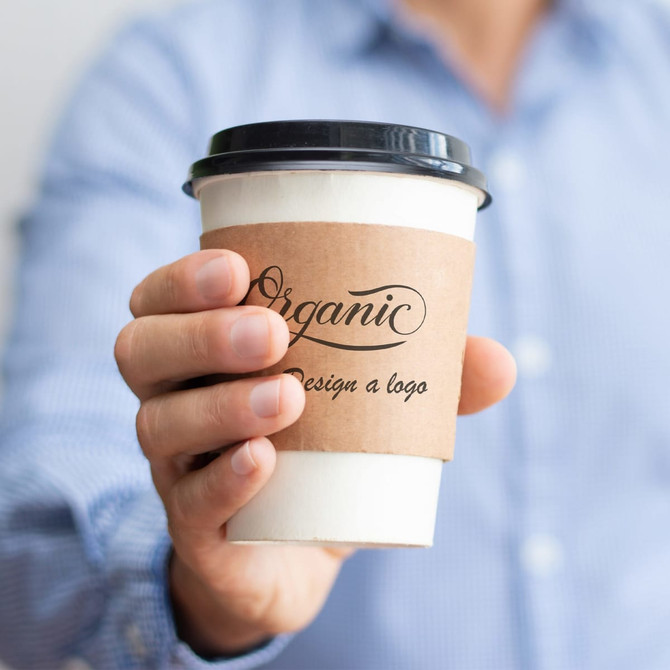 A drink cup with a black lid and a brown paper sleeve, held by a person in a blue shirt.