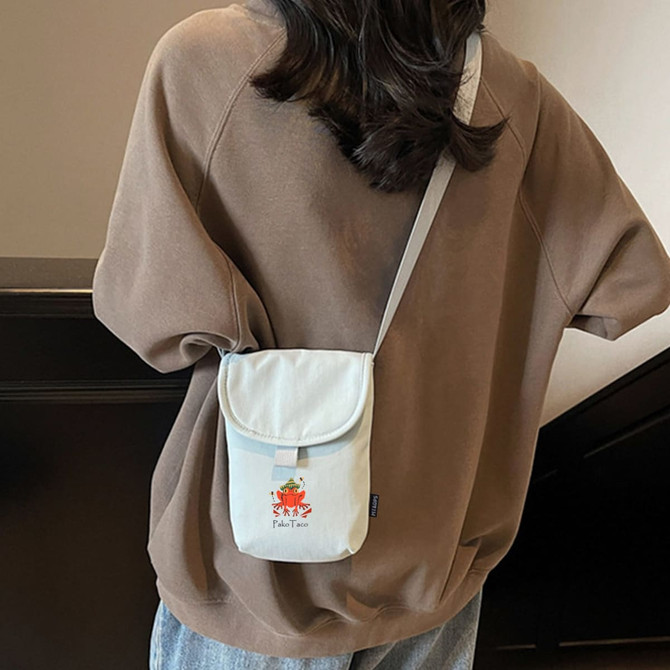 A small canvas shoulder bag in light grey with a flap, carried on a person's shoulder, wearing a brown oversized top.