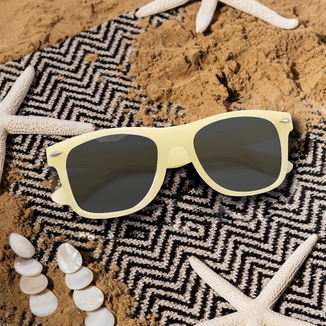 Glow in the dark sunglasses in pale yellow, resting on a patterned beach mat with seashells and starfish.