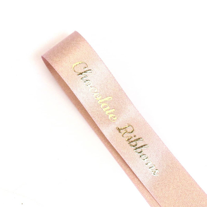 A satin ribbon in a soft pink shade with a metallic gold inscription, featuring a logo.