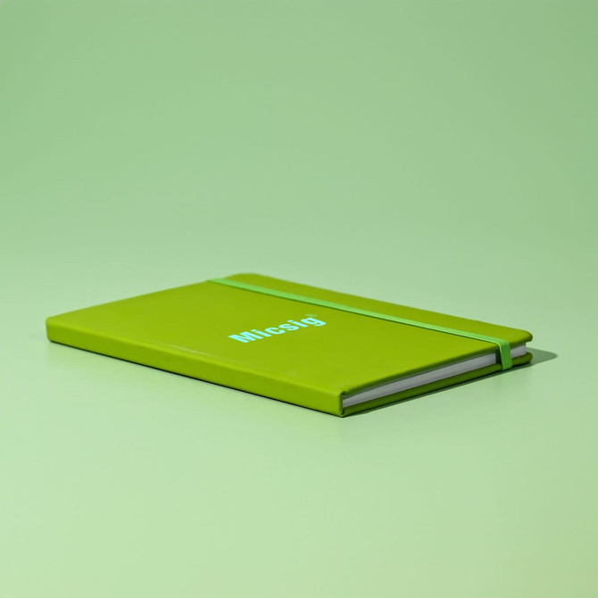A green A5 notebook with an elastic band and a simple logo on the cover, resting on a matching green surface.
