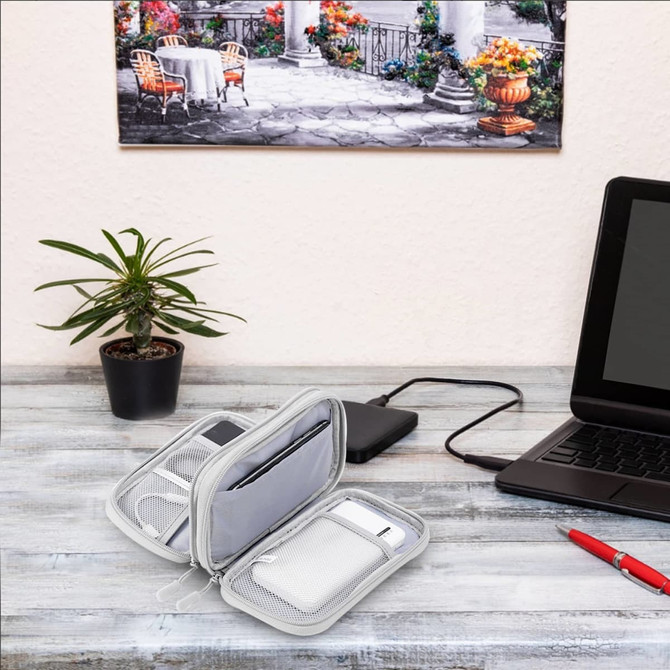 A grey electronic accessory organiser bag with multiple compartments, displayed on a desk alongside a laptop and a plant.
