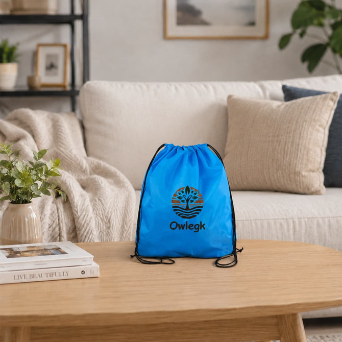 A blue drawstring bag sits on a wooden table in a cosy living room setting with a plant nearby. It features a logo.