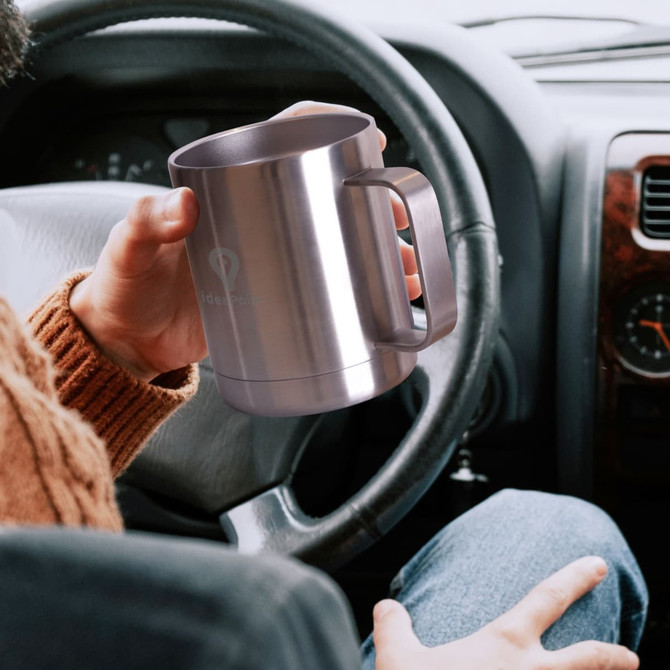 A stainless steel coffee mug with a handle, held in a hand inside a car. The mug has a logo on its side. A stainless steel coffee mug with a handle, held in a hand inside a car. The mug has a logo on its side.
