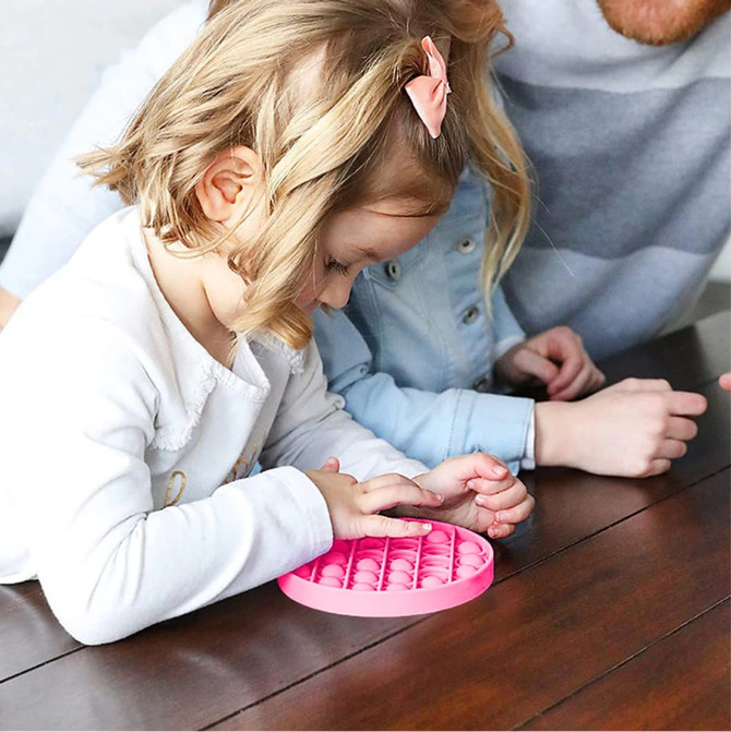 A pink Pop It fidget toy rests on a wooden table, while a child interacts with it, alongside another person.