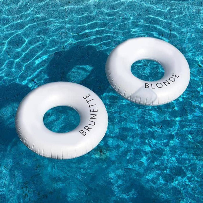 Two white inflatable swim rings floating in a blue pool, featuring the words "BRUNETTE" and "BLONDE."