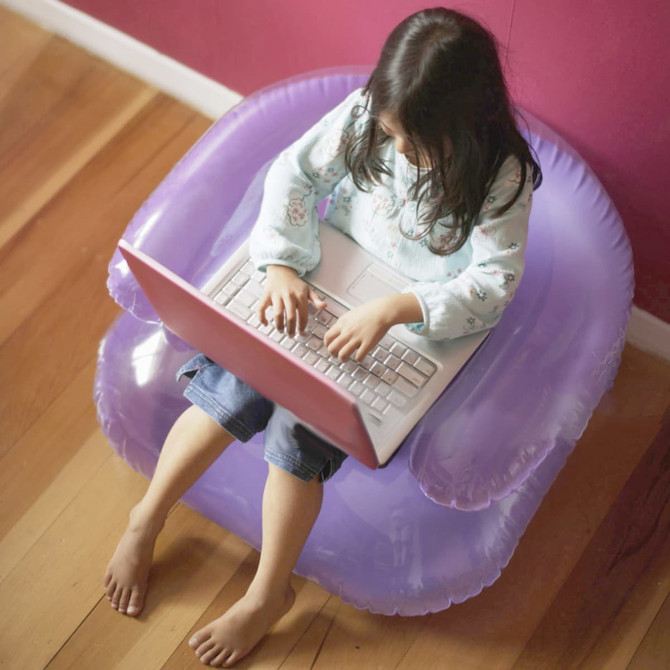 A purple inflatable chair with a child sitting on it, using a laptop. The background features a pink wall.