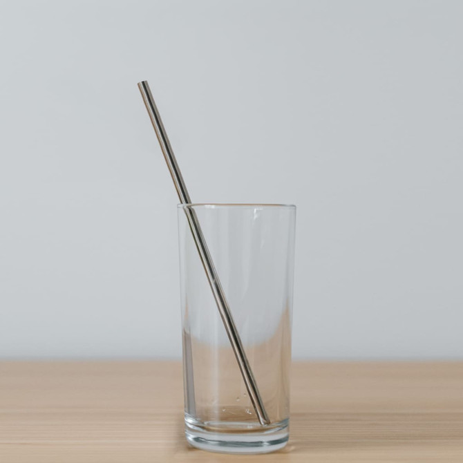 A drinking glass containing a stainless steel straw, placed on a wooden surface against a light background.