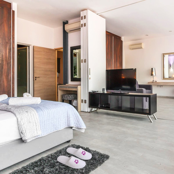 A pair of white hotel slippers placed on a grey rug in a modern hotel room with wooden furniture and a TV.