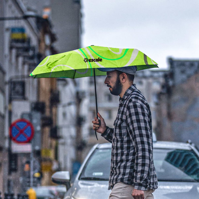 A green mini folding umbrella with a case, held by a person in a checked shirt while walking in the rain.
