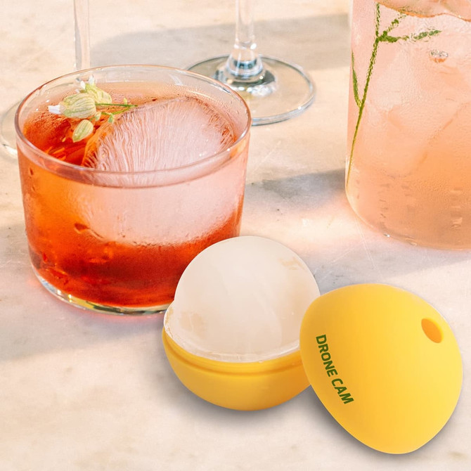 A yellow silicone whisky ice ball mould next to a glass of cocktail featuring an ice ball and garnished drink.