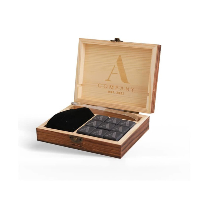 A wooden box containing grey whiskey stones, a pair of tongs, and a black storage pouch, featuring a logo on the lid.