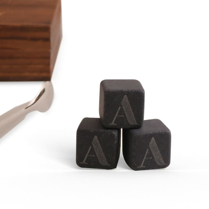 A set of three black whiskey stones with engraved letters, alongside a metal tongs and a wooden box.