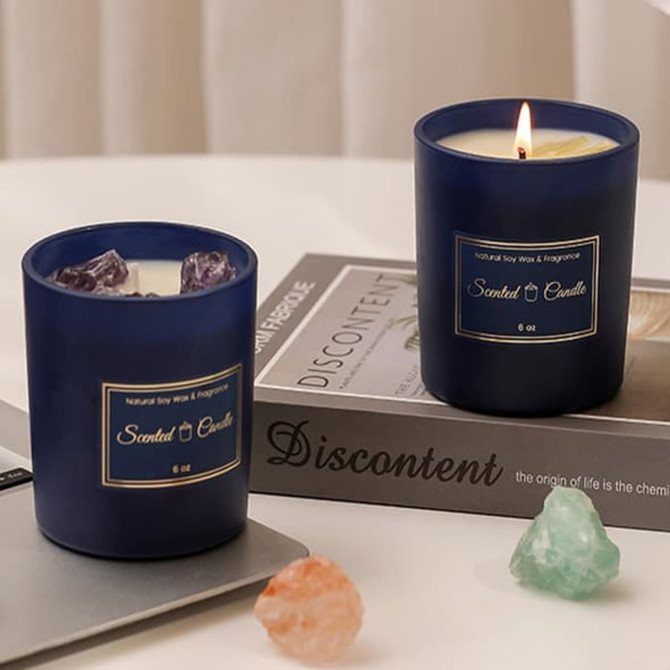 Two navy glass candles with a glowing flame on one, surrounded by crystals and placed on a decorative book.