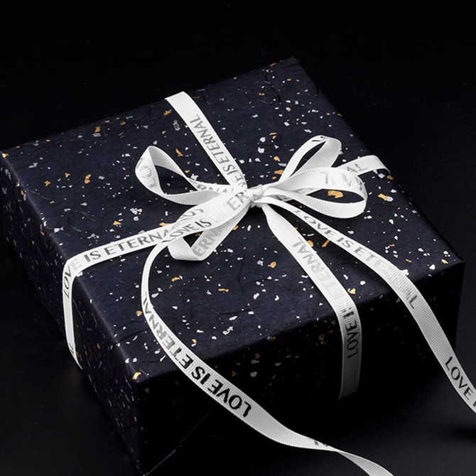 A wrapped gift box in dark blue with a white ribbed fabric ribbon and a logo. The ribbon features printed text.