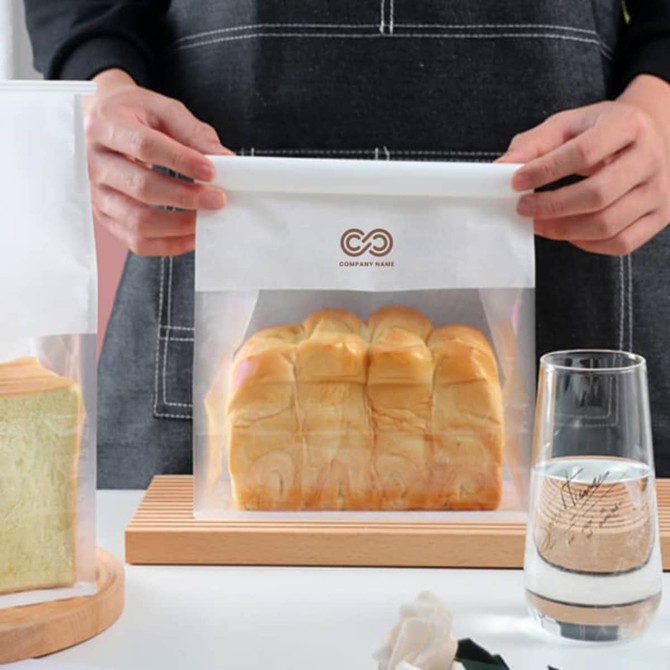 A person holds a clear, resealable bag containing sliced bread. The bag features a logo at the top. A person holds a clear, resealable bag containing sliced bread. The bag features a logo at the top.