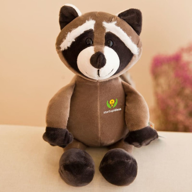 A brown raccoon plush toy with a white face and black markings, featuring a logo on its chest.