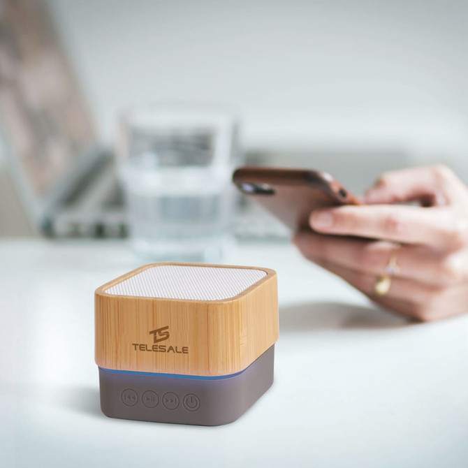 A bamboo Bluetooth wireless speaker with grey base, placed on a table beside a laptop and a drink.