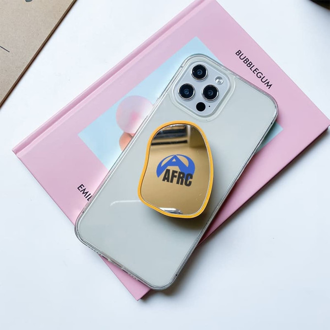 A yellow custom-shaped mirror phone pop holder on a clear phone case, resting on a pink book.