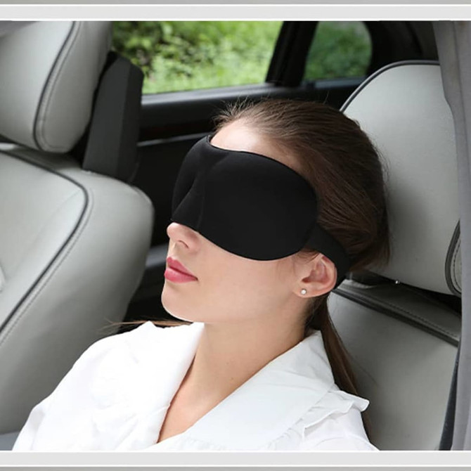 A cushioned black eye mask resting on a woman’s face, situated in a car seat. It has adjustable straps.
