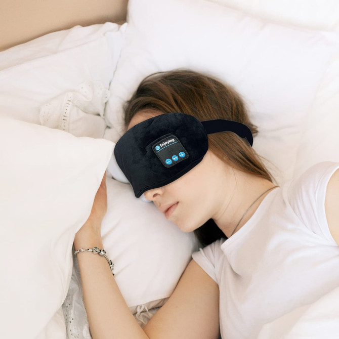 A pair of black Bluetooth sleep headphones worn by a person resting on a bed with white pillows.