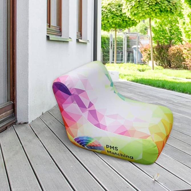 An inflatable lounge chair in a rainbow gradient design, placed on a wooden deck outside. It has a logo on the side.