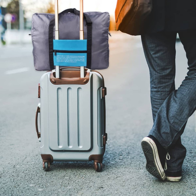 A grey travel compressed bag attached to a silver suitcase, being wheeled along a pavement by a person in dark clothing.