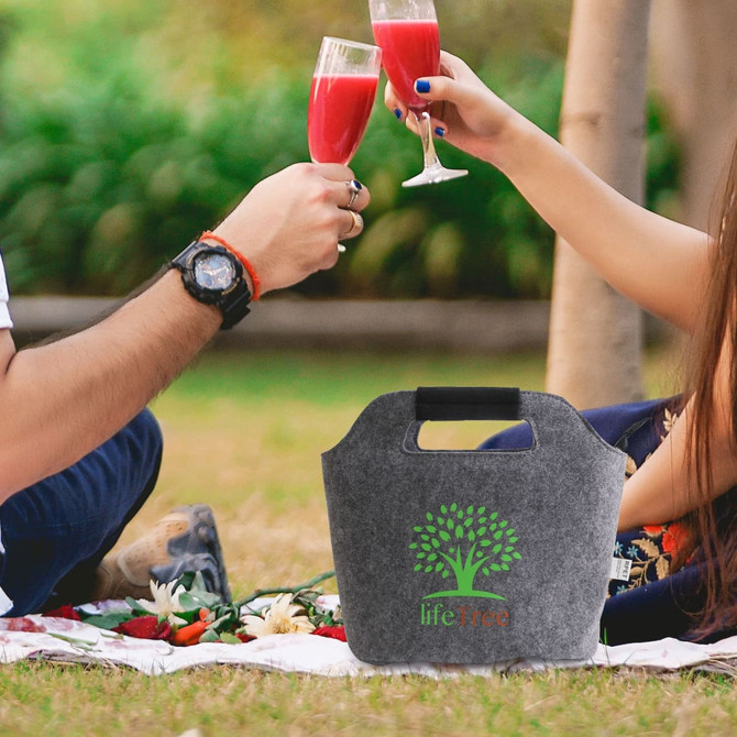 A grey felt cooler bag with a handle and logo, set on a picnic blanket, surrounded by drinks and greenery.