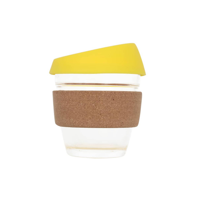 A reusable glass coffee cup with a cork band and a yellow silicone lid. The cup is clear.