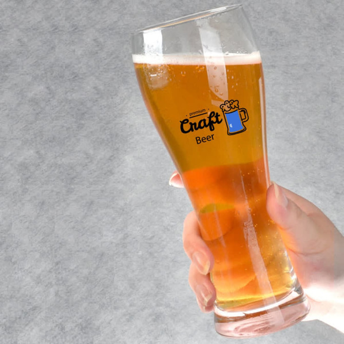 A tall, clear pint beer glass filled with amber beer, held by a hand. The glass features a logo.