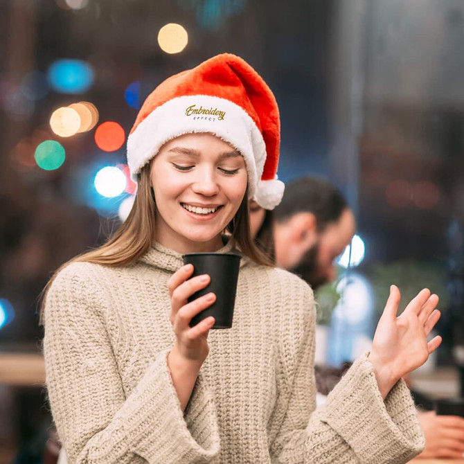 A red and white plush Santa hat worn by a smiling woman holding a black cup, with blurred festive lights in the background.