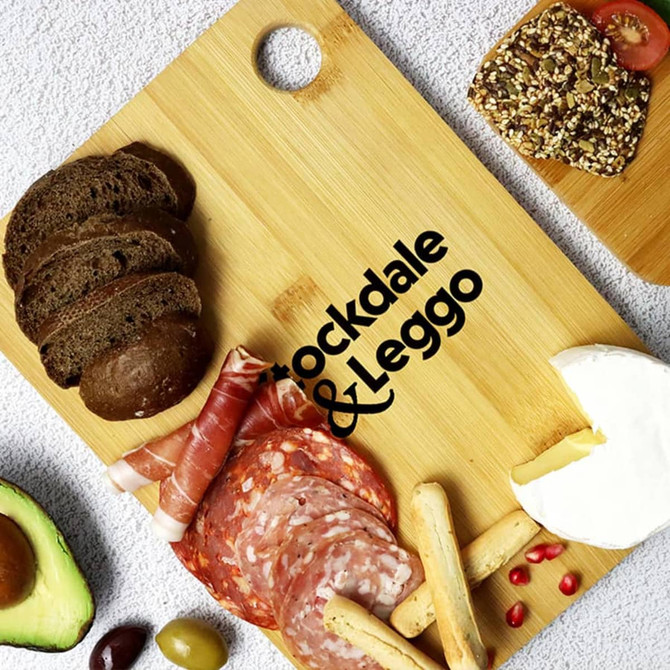 A large bamboo cutting board with assorted food items, including bread, meats, cheese, and an avocado, featuring a logo.