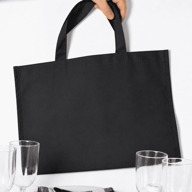 A black picnic backpack with sturdy handles, placed near glasses and tableware. It has a simple, minimalistic design.