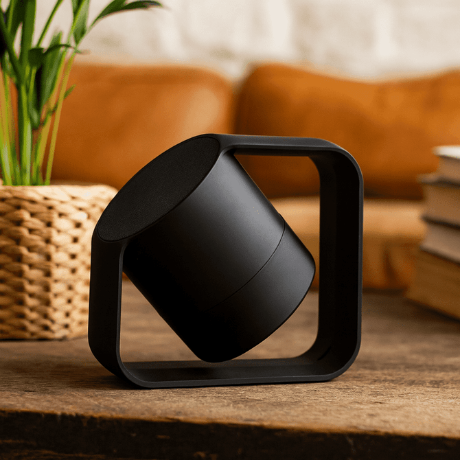 A black wireless speaker with a unique geometric design, set against a decorative background with plants and books.