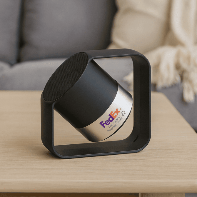 A wireless speaker in silver and black, featuring a square frame and a logo. Made from recycled ABS and aluminium.