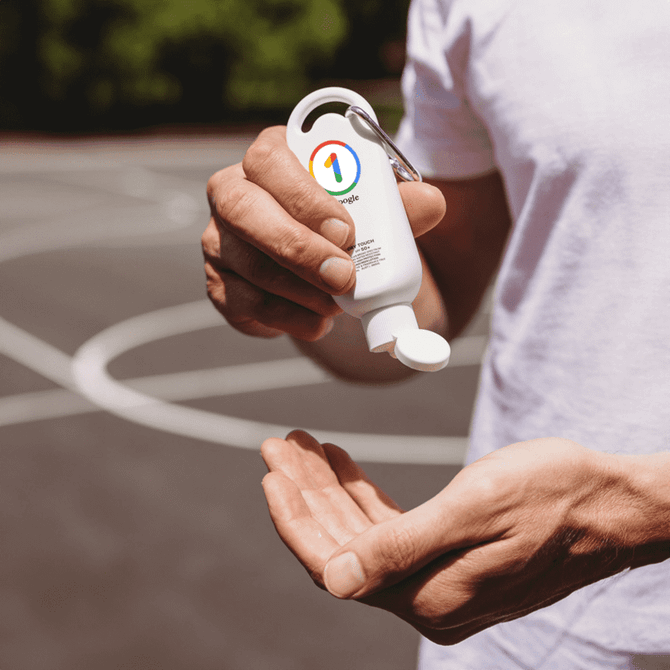 A white sunscreen bottle with a keychain cap, held above a hand, set against a blurred outdoor background.