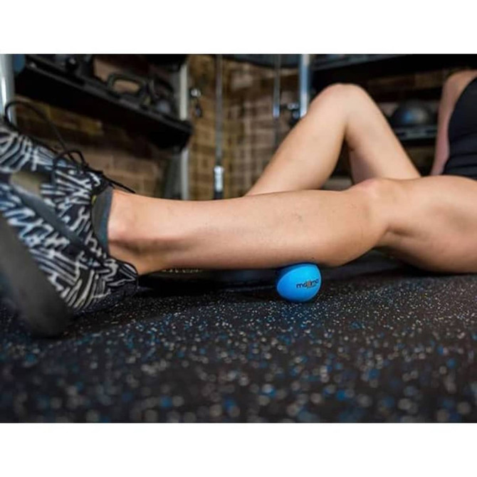 A blue massage ball sits on the floor alongside a person's leg, set against a gym backdrop.