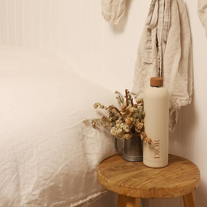 A drink bottle in a soft beige colour with a wooden lid, featuring a logo, sits on a wooden stool beside dried flowers.