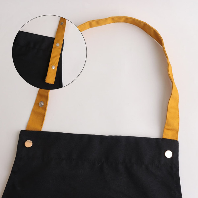 A black poly-cotton canvas apron featuring a yellow colour neck strap and metal snap buttons.