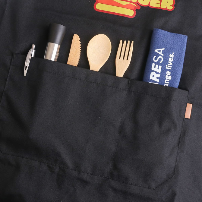 A black canvas apron featuring a pocket with wooden utensils, a pen, and a blue item with a logo.