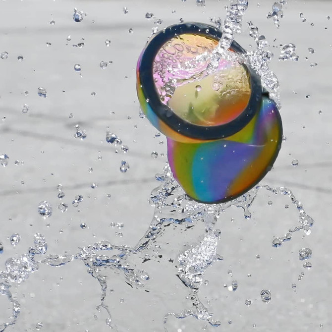 A colourful, reusable water balloon splashes through water droplets against a grey background.