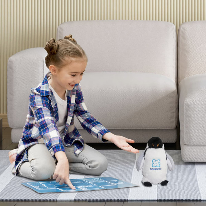 A child sits on a rug, reaching toward a cute grey and white penguin plush toy, with a logo on its front.