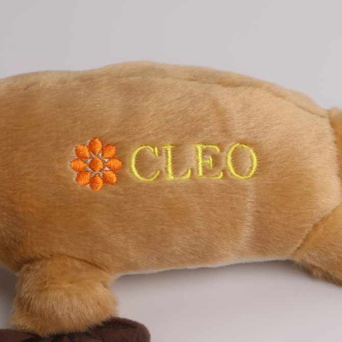 A soft, plush toy in tan, shaped like a platypus, with an embroidered logo in orange and yellow.