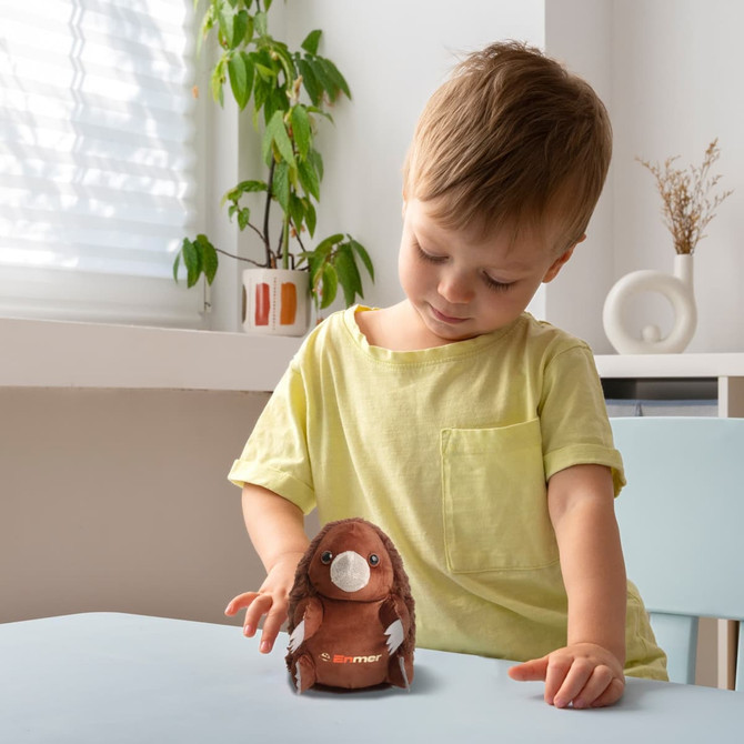A child in a light yellow shirt holds a brown echidna plush toy on a table, with plants in the background.