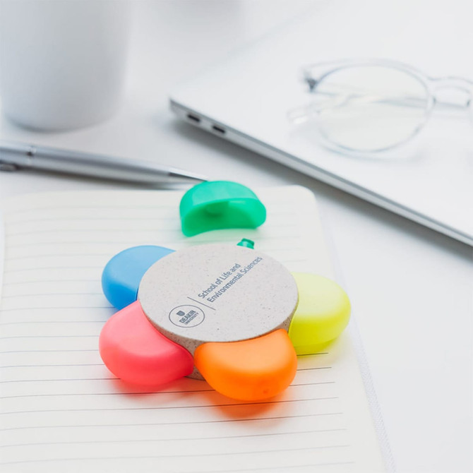 A flower-shaped set of highlighters in pink, orange, yellow, green, and blue, on a notebook with a pen and glasses nearby. A flower-shaped set of highlighters in pink, orange, yellow, green, and blue, on a notebook with a pen and glasses nearby.